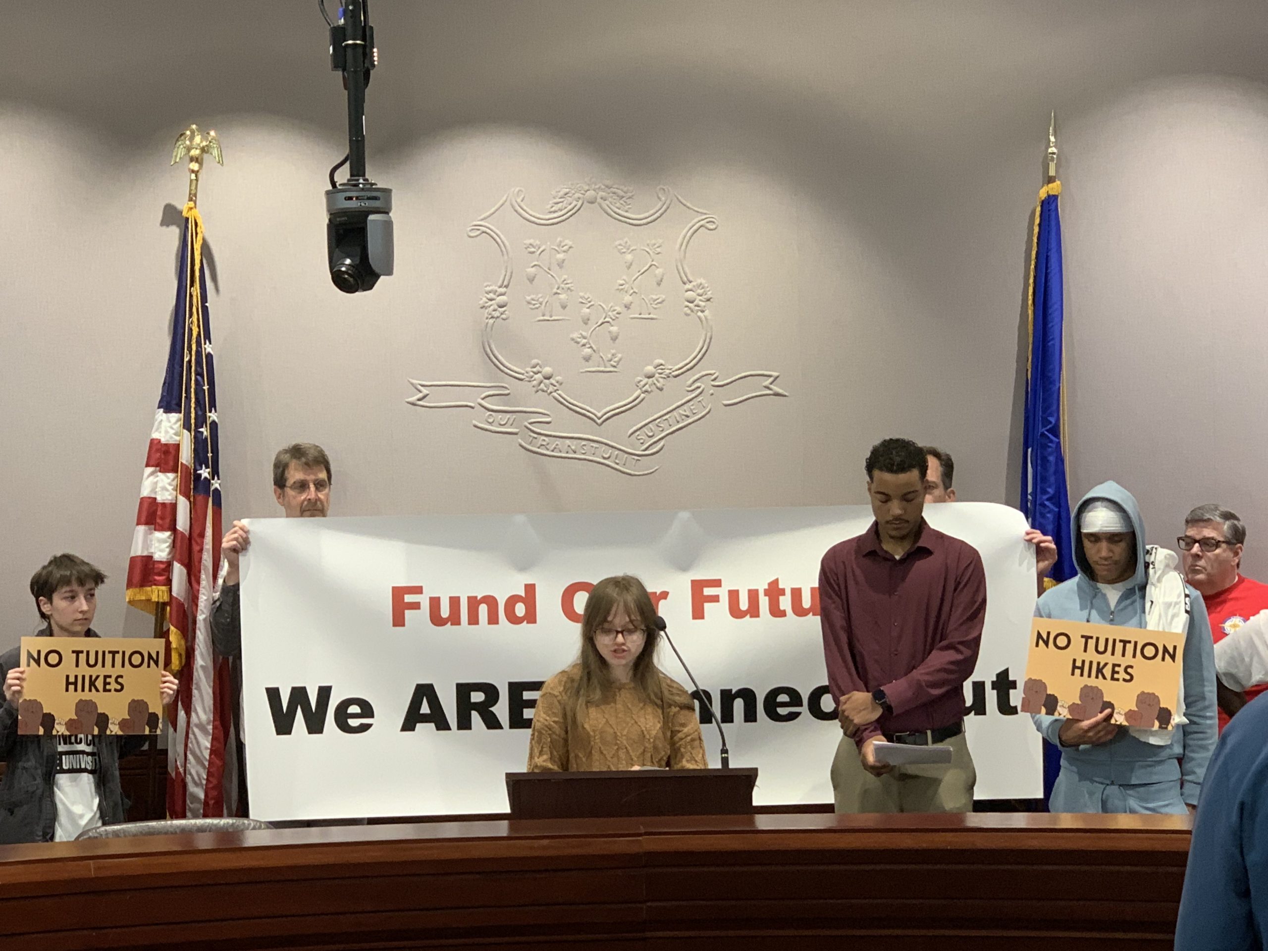 A CCSU student standing at a press conference in front of students holding signs reading “fund our future, we are Connecticut” and “no tuition hikes”
